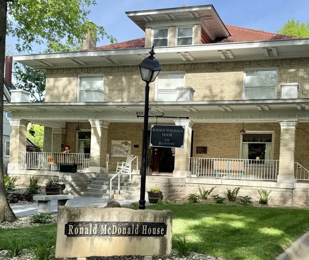 The exterior of the Ronald McDonald House of Northeast Kansas, featuring a large multi-story home with a covered front porch surrounded by green trees.